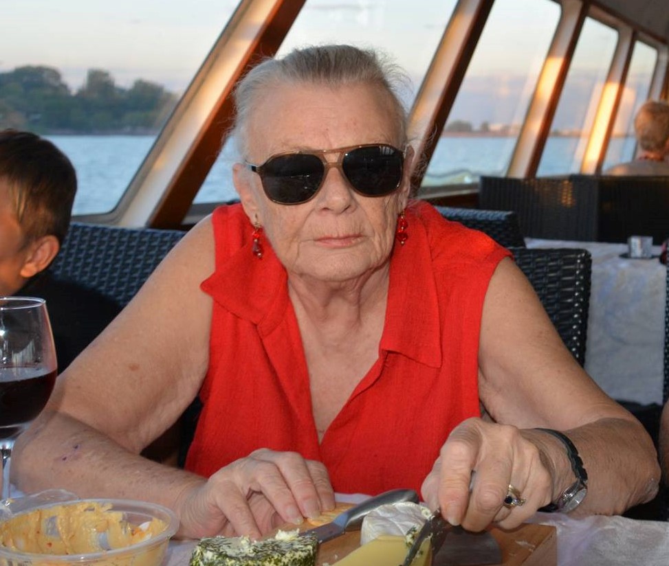 Image of Rhonda, a woman who looks to be in her seventies, wearing a red top, sunglasses some earings and is seen holding a cheese knife, with some cheeses, a glass of wine and dip.